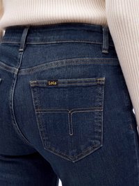 Close-up af mørkeblå denim jeans baglomme med gul syning og lille "Lois" label, båret med en beige ribstrikket sweater.