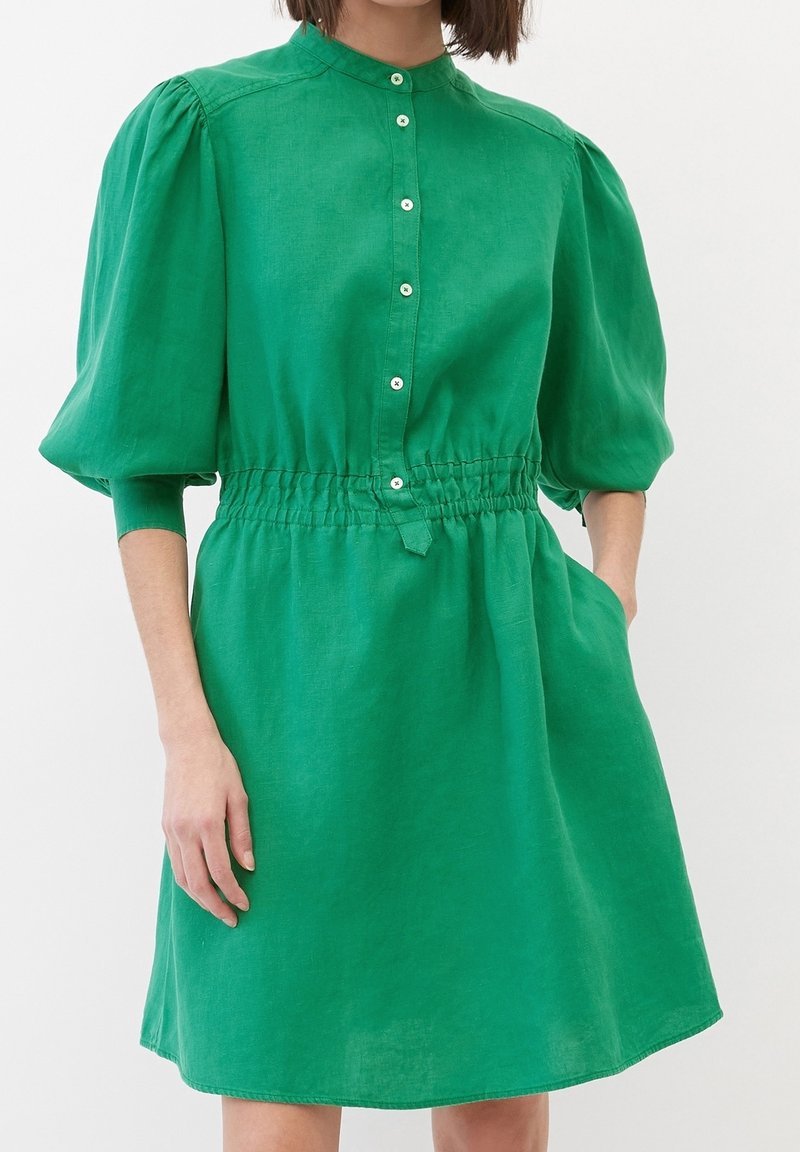 Donna che indossa un vestito verde brillante con maniche a sbuffo lunghe fino al gomito, davanti con bottoni, vita elasticizzata e tasche laterali.