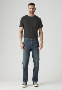 Vyras stovi vilkėdamas juodą marškinėlį, mėlynus džinsus ir juodus sportbačius su baltais padais prieš neutralų foną.