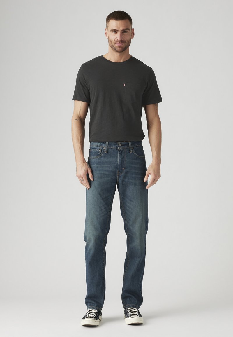 Vyras stovi vilkėdamas juodą marškinėlį, mėlynus džinsus ir juodus sportbačius su baltais padais prieš neutralų foną.