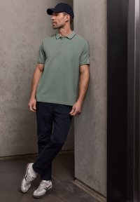 Homme portant un polo vert, un pantalon bleu marine, des baskets blanches et une casquette noire, appuyé contre un mur en béton à l'intérieur.