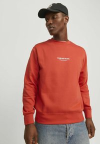 Oranje sweatshirt met ronde hals en geribbelde manchetten, voorzien van een wit "Originals Studio" logo op de borst, gedragen met lichtblauwe jeans.