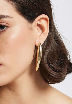 Goldene Creolen mit einer glatten, glänzenden Oberfläche, länglich ovaler Form und dünnem Design, die am Ohrläppchen gegen dunkles Haar getragen werden.