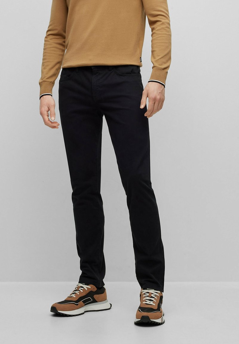 Jeans negros de corte slim hechos de algodón con una textura suave. Combinados con un suéter color camel y zapatillas con detalles marrones y cordones blancos.