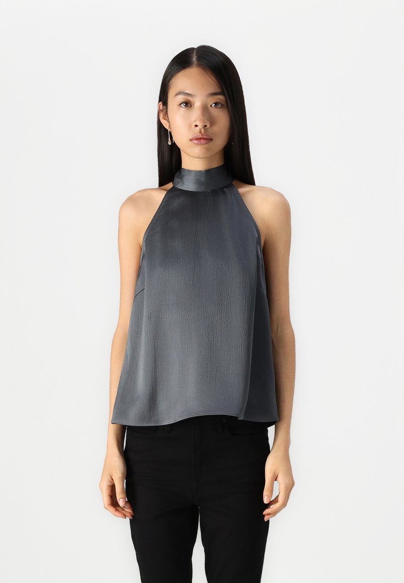 Haut sans manches en halter gris foncé, avec un tissu texturé à finition lisse, un design drapé et un col montant. Associé à un pantalon noir.