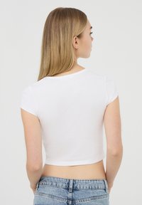 T-shirt bianco cropped con maniche corte, realizzato in tessuto leggero. Presenta un colletto rotondo e una texture liscia, abbinato a jeans in denim blu.