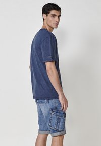 Temno modra bombažna majica s sproščenim krojem, v kombinaciji s svetlo modrimi denim hlačami, ki imajo več žepov in zavite roba.