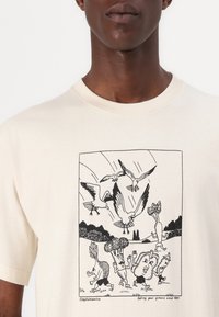 Beige bomulds T-shirt med en sort grafisk illustration af animerede fugle og grøntsager i humoristiske positurer, indrammet med en rektangulær kant.
