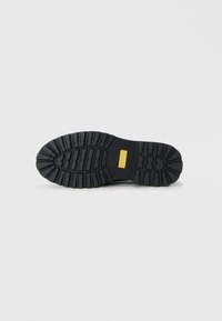 Suela de goma negra con surcos profundos para agarre, con un acento amarillo en la sección plana, diseñada para durabilidad y tracción.