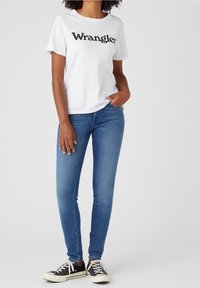 Bílá bavlněná tričko s potiskem "Wrangler" v černé barvě. Kombinováno s úzkými modrými džínami a černými teniskami s bílou podešví.