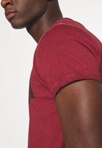 Camiseta de manga corta color burdeos hecha de algodón suave. Presenta un cuello redondo y los bordes de las mangas ligeramente desgastados. Textura suave y casual.