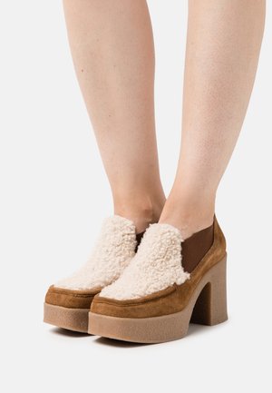 Chaussures compensées en daim marron avec une tige blanche duveteuse. Caractérisées par un talon épais et un bout arrondi. Conçues pour un enfilage facile avec une finition texturée.