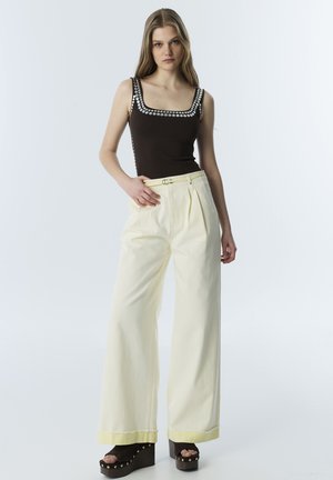 Vrouw staand met hand op heup, draagt zwart mouwloos topje met zilveren studs, brede crèmekleurige broek en platformsandalen met studs.