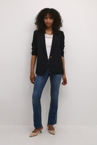 Žena s kudrnatými vlasy má na sobě černé sako přes bílý krajkový top, modré džíny a béžové boty s nízkým podpatkem, stojí před jednobarevnou bílou zdí.