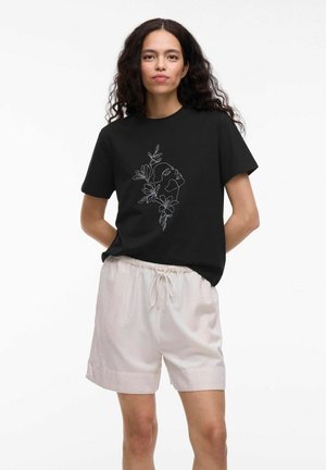 T-shirt nera con disegno bianco di un volto e foglie sul davanti, abbinata a pantaloni corti beige chiaro con vita regolabile con cordino.