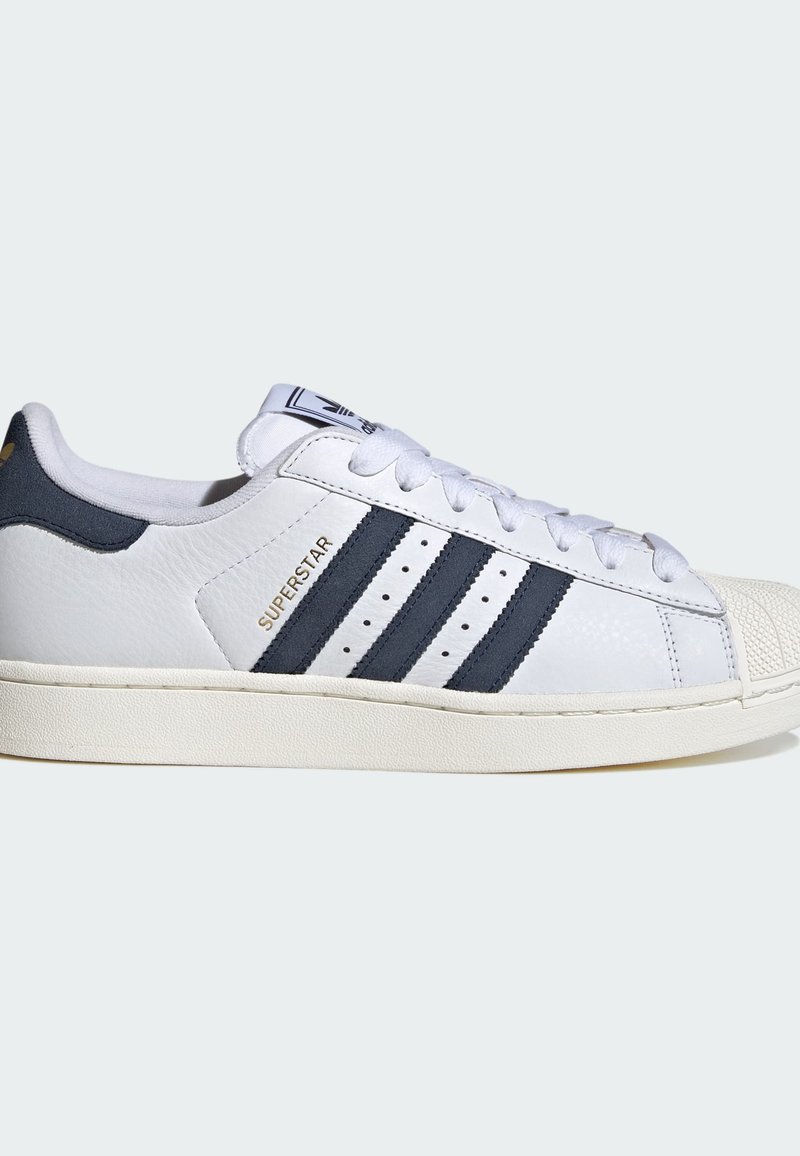 White Adidas Superstar sneaker with black stripes, rubber shell toe, and "SUPERSTAR" text in gold on side, shown in right profile.