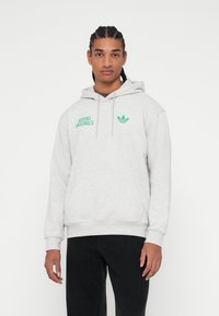 Szary pulower z kapturem wykonany z miękkiego materiału, z kieszenią typu kangur, kapturem ze sznurkiem oraz zielonym logo "Adidas Originals" na piersi.