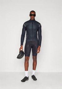 Černá cyklistická bunda s dlouhými rukávy, celým zipem a reflexními detaily, v kombinaci s černými cyklistickými kraťasy a botami, držící černou helmu.