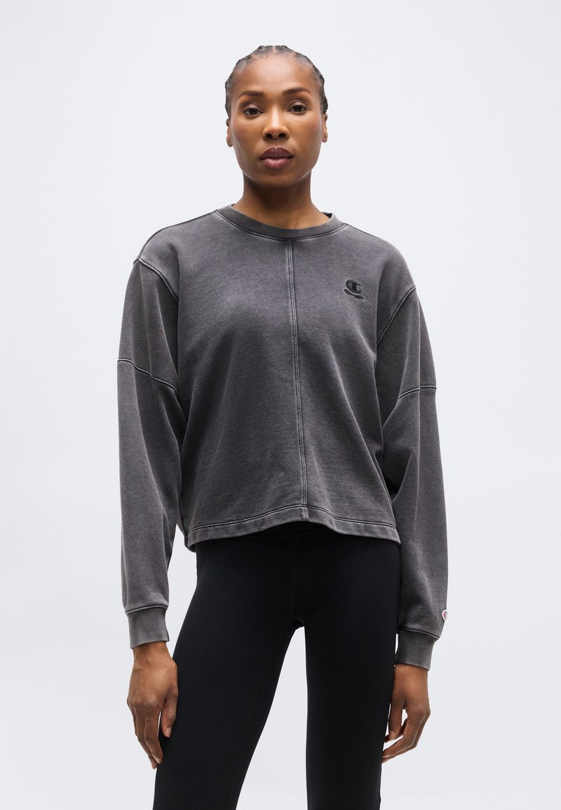 Γυναίκα με πλεξούδες, που φοράει σκούρα γκρι φούτερ Champion και μαύρα κολάν, στέκεται μπροστά σε απλό φωτεινό φόντο.