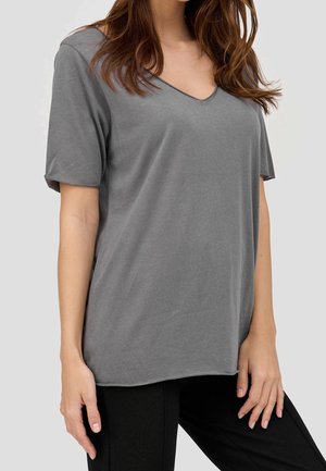 Femme portant un t-shirt ample gris à col en V et manches courtes, et un pantalon noir, cheveux lâchés, debout devant un fond clair uni.