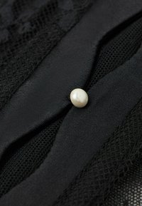 Sort stof med spidse og teksturerede mesh-lag, prydet af en enkelt oval hvid knap i midten. Glatte og indviklede detaljer.