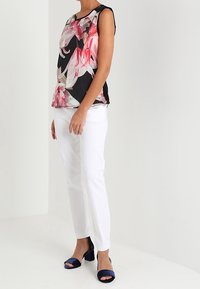 Top sans manches à motif floral noir avec des accents roses et verts, associé à un pantalon blanc et des sandales à talons hauts bleu marine.