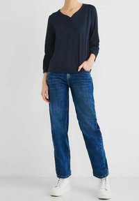 Marinblå långärmad topp med V-ringning och kort dragkedjedetalj, kombinerad med löst sittande blå jeans och vita sneakers.