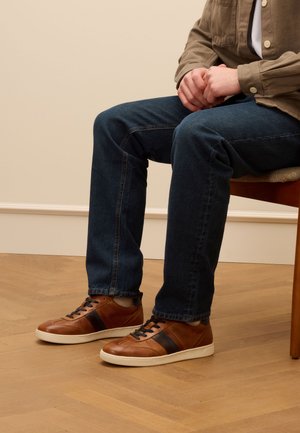 Sneakers in pelle marrone con strisce nere, pannelli testurizzati e suole in gomma bianca, abbinate a jeans scuri, seduto su una sedia di legno.