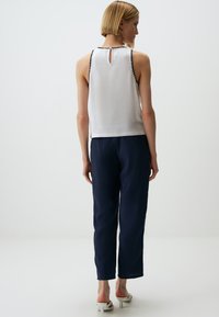 Witte mouwloze top met zwarte bies, met een keyhole rug, gecombineerd met marineblauwe broek en witte block-heeled sandalen.