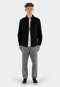 Sort tekstureret jakke med knaplukning, to brystlommer, båret over en hvid t-shirt, parret med grå jeans og sorte sneakers.