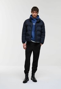 Jachetă puffer bleumarin cu un design matlasat, purtată peste un hanorac albastru. Pantaloni negri tapered și botine strălucitoare completează ținuta.