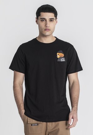 Homme portant un t-shirt noir avec un motif d'ours en peluche sur la poche poitrine et un pantalon beige avec une étiquette sur la poche.
