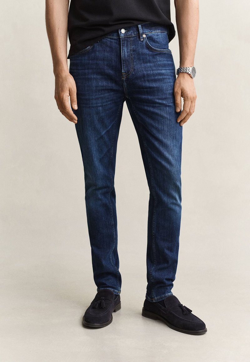 Homme portant un jean slim bleu foncé, des mocassins noirs à pampilles, une chemise noire et une montre-bracelet argentée au poignet gauche.