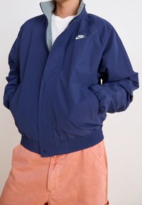 Mörkblå vindjacka med en ljusgrå krage, ribbade ärmslut och midja. Har en liten vit Nike-logotyp på br östet.