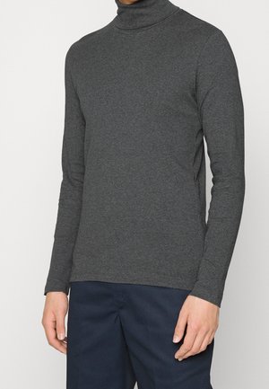 Mand iført en tætsiddende, langærmet, mørkegrå rullekravesweater kombineret med mørkeblå bukser, stående foran en ensfarvet baggrund.