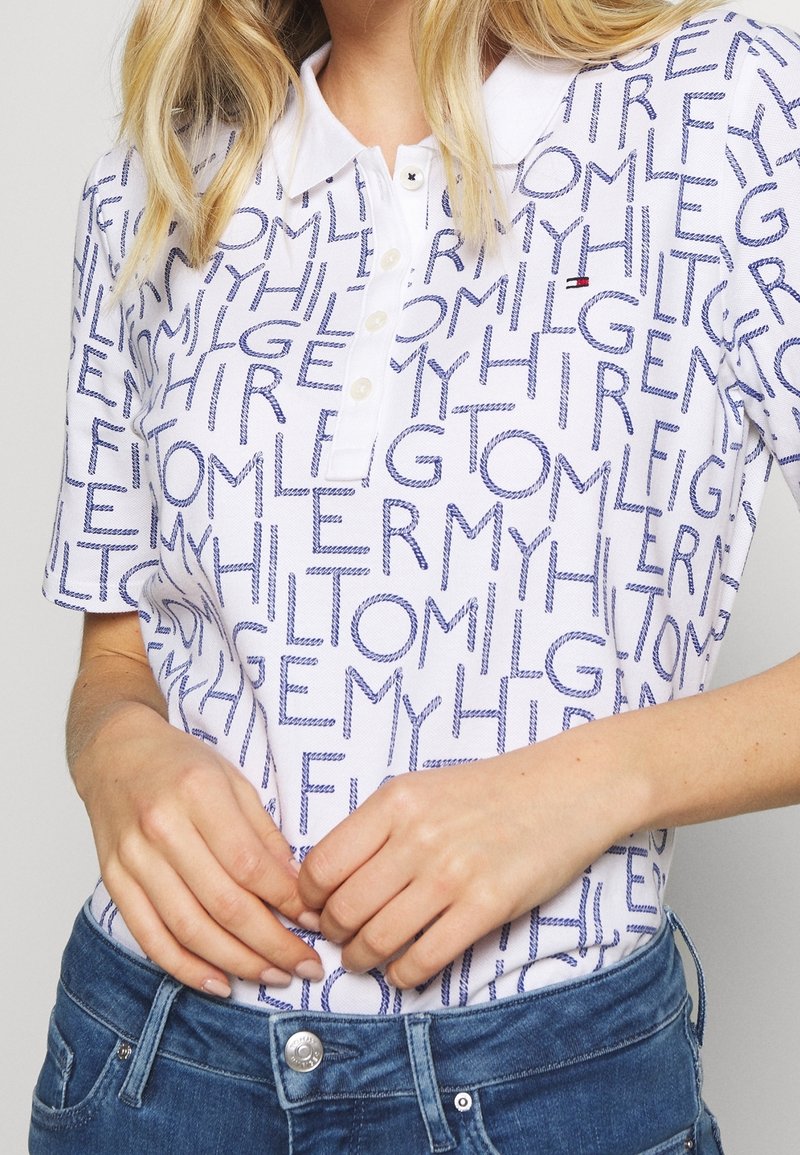 Femme portant un polo blanc avec un motif de texte bleu et un jean bleu, mains jointes près de la taille.