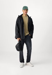 Chaqueta impermeable negra, camisa caqui, jeans oscuros, zapatillas blancas y una gorra verde. Sostiene un bolso negro con una marca blanca.