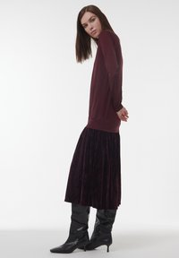 Frau mit glattem braunem Haar, die ein langärmliges bordeauxfarbenes Oberteil, einen passenden Samtenrock und schwarze Stiefel mit Absatz trägt, vor einem weißen Hintergrund.