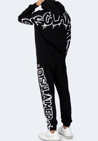 Persona che indossa un hoodie nero e pantaloni da tuta con grande testo in stile graffiti "DISCLAIMER" in bianco, abbinati a sneaker bianche.