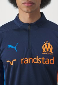 Tamsiai mėlynas sportinis puloveris su aukšta apykakle ir ketvirtį užtrauktuku. Puikuojasi oranžinėmis logotipais: "randstad" ir Marselio emblema, su mėlynu Puma logotipu.