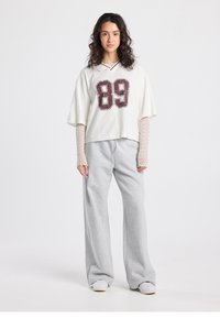 T-shirt blanc court avec impression rouge "89", associé à un pantalon de survêtement ample gris clair. Manches longues texturées avec un motif en résille.
