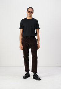 Svart t-shirt med rund hals, bruna kordflorsbyxor och svarta snörskor. Modellen har på sig solglasögon och står mot en enfärgad bakgrund.