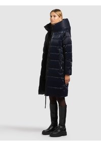 Žena stoji u crnom podstavljenom dugom kaputu s visokim ovratnikom, crnim tajicama i crnim debelim čizmama do koljena na jednobojnoj svijetloj pozadini.