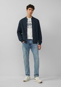 Marine bomberjack met een ronde kraag, knoopsluiting, witte T-shirt met opdruk, lichtblauwe jeans en witte sneakers met beige accenten.