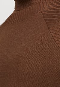 Pull marron avec un col montant côtelé, un tissu texturé et un motif tricoté lisse avec un léger éclat. Gros plan sur les détails de l'épaule.