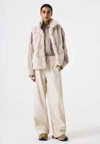 Chaleco de piel sintética crema, camiseta de rayas beige y pantalones sueltos beige. El calzado es beige con acentos oscuros. Textura simple y suave en todo.