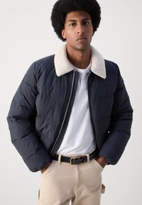 Pufjakke i mørk marineblå med en krave i creme-faux shearling, har lynlås-lukning og quiltet tekstur, båret over en hvid skjorte.