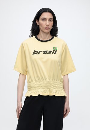 Kobieta z długimi ciemnymi włosami ubrana w żółtą bluzkę z krótkim rękawem z czarnym napisem "Brasil", brazylijską flagą i czarnymi spodniami na jednolitym tle.