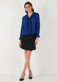 Blue blouse with ruffled neckline and long sleeves, paired with a black skirt. The blouse features button details and a smooth texture.