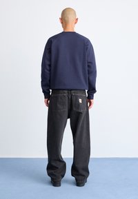 Persona con cabeza rapada vestida con sudadera navy y jeans negros sueltos, de pie descalzo sobre un suelo azul frente a una pared blanca, de espaldas a la cámara.